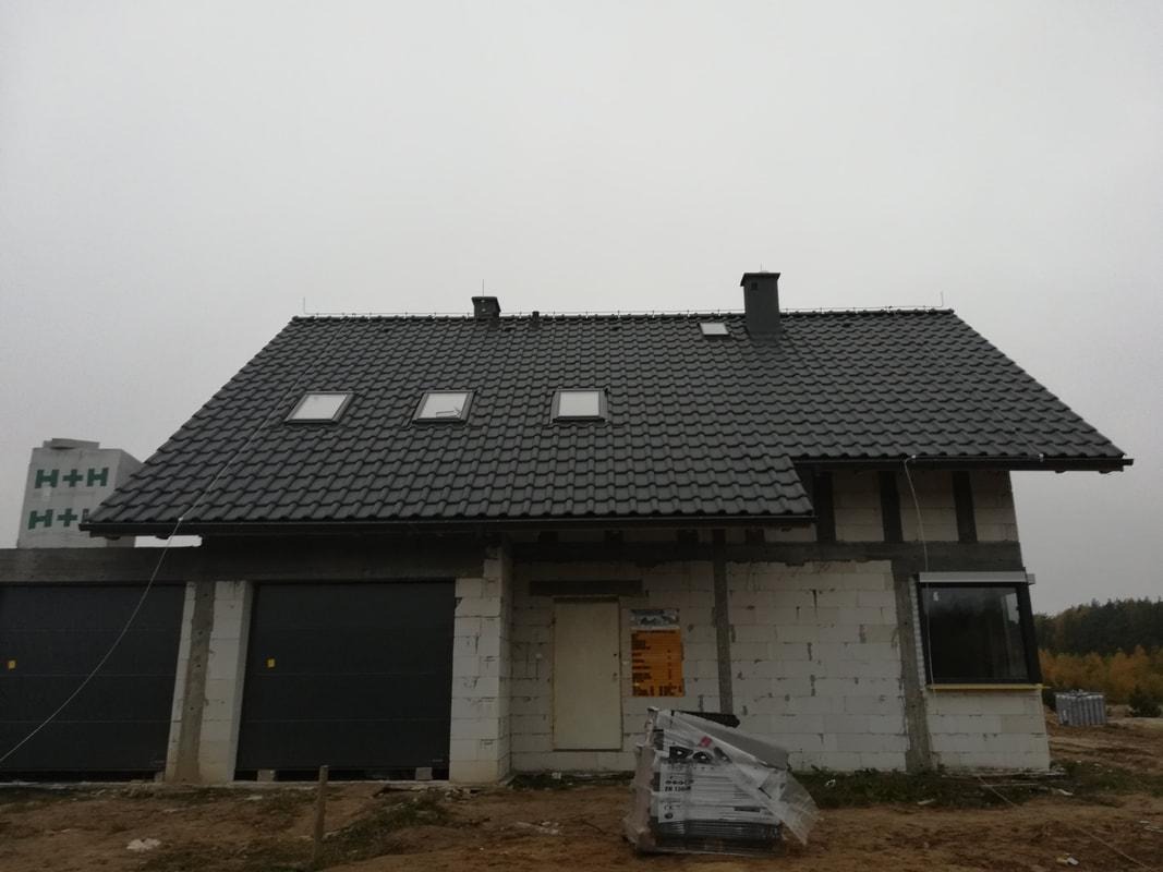 Dom w trakcie budowy z nowym, ciemnoszarym dachem z dachówką, trzema oknami dachowymi i kominem, widoczne elementy konstrukcyjne budynku z betonu komórkowego oraz pakiet dachówek na ziemi.