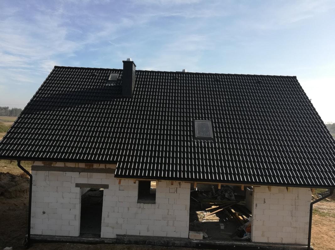 Dom w stanie surowym otwartym z czarną dachówką karpiówką, widoczne okna dachowe i orynnowanie, w tle błękitne niebo.