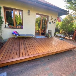 ARTUR MAJKA 3M3 - Nowoczesny, drewniany taras przydomowy z leżakiem i widokiem na zadbany ogród, wykonany z desek o ciepłym, brązowym odcieniu, ułożonych równolegle do ściany domu.