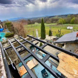Konstrukcja z czarnych belek na kamiennej podstawie, prawdopodobnie w trakcie budowy zadaszenia tarasu, z widokiem na rozległy krajobraz i pochmurne niebo.