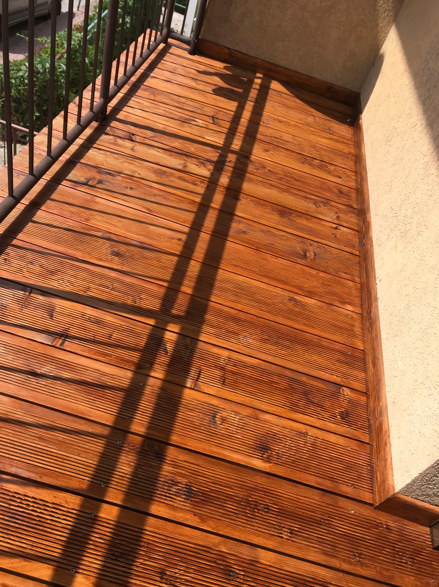 Nowy, lśniący taras z desek w odcieniach brązu, z widocznymi śladami impregnacji, oświetlony promieniami słonecznymi tworzącymi cienie balustrady.