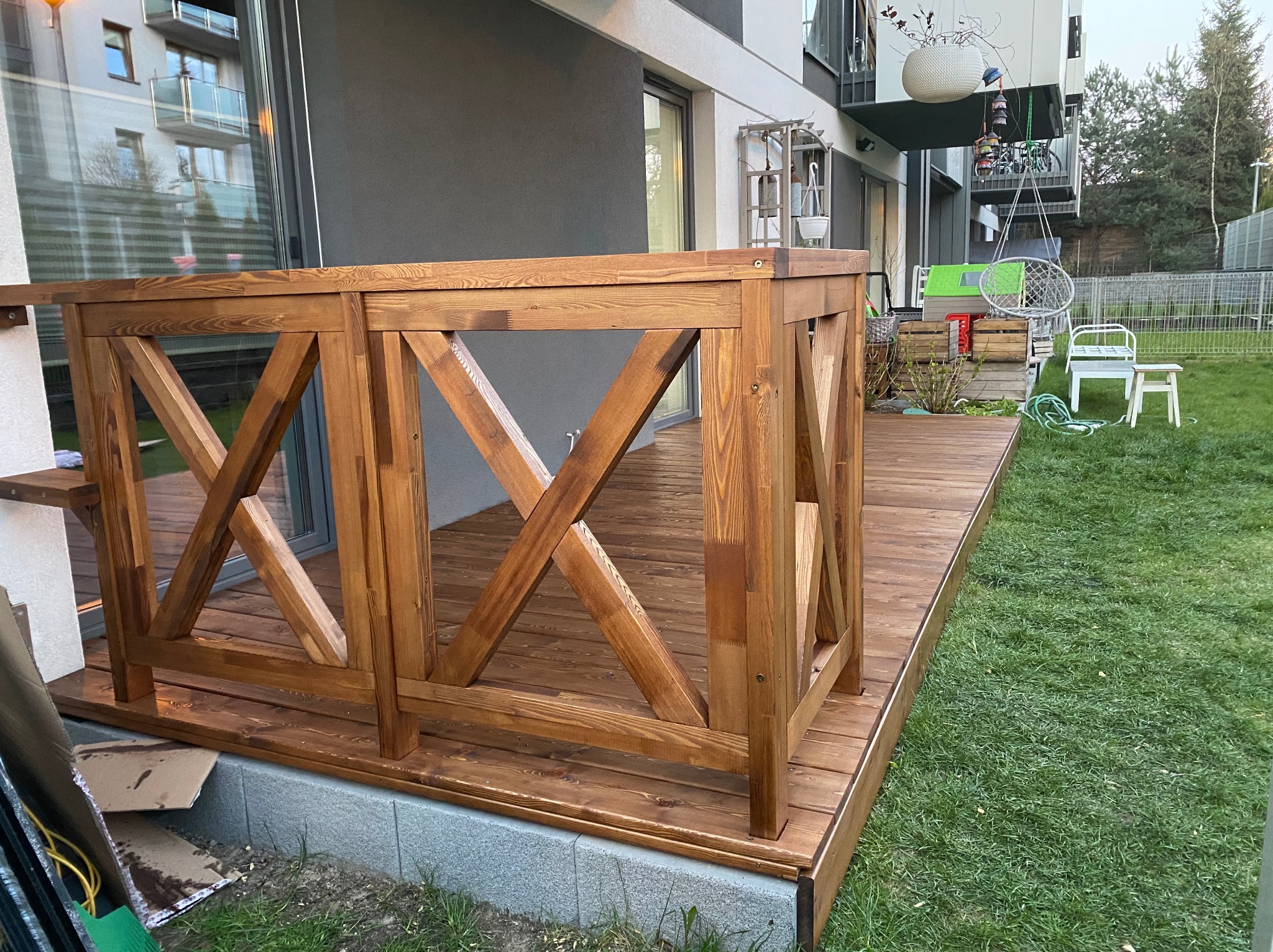 Nowoczesny taras drewniany z balustradą w kształcie krzyża, świeżo po impregnacji, z widoczną strukturą drewna i elementami montażowymi, otoczony zielonym trawnikiem.