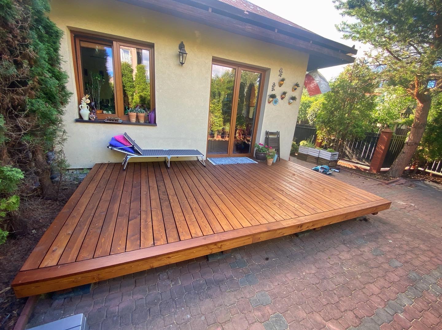 Nowoczesny, drewniany taras przydomowy z leżakiem i widokiem na zadbany ogród, wykonany z desek o ciepłym, brązowym odcieniu, ułożonych równolegle do ściany domu.