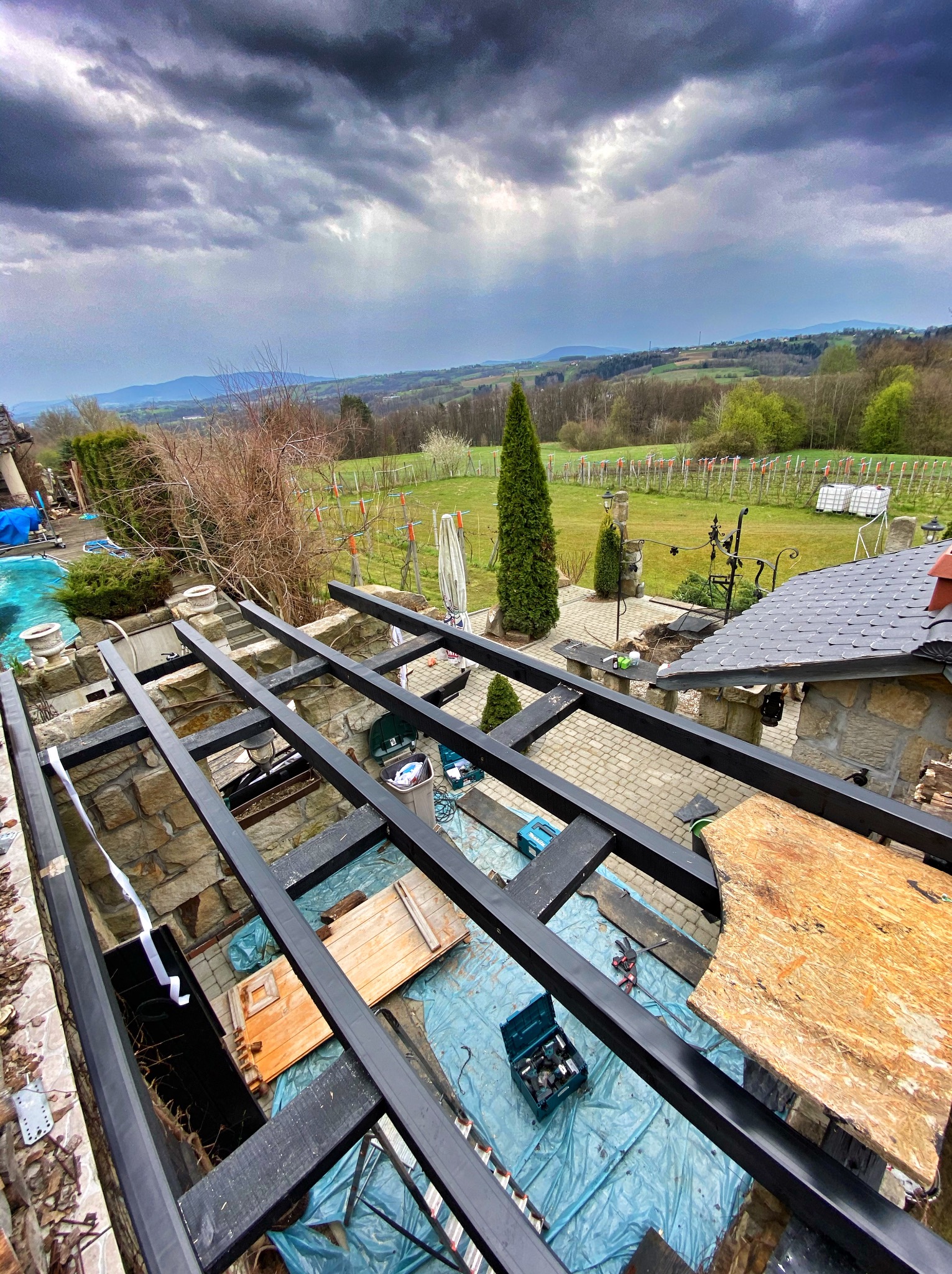 Konstrukcja z czarnych belek na kamiennej podstawie, prawdopodobnie w trakcie budowy zadaszenia tarasu, z widokiem na rozległy krajobraz i pochmurne niebo.