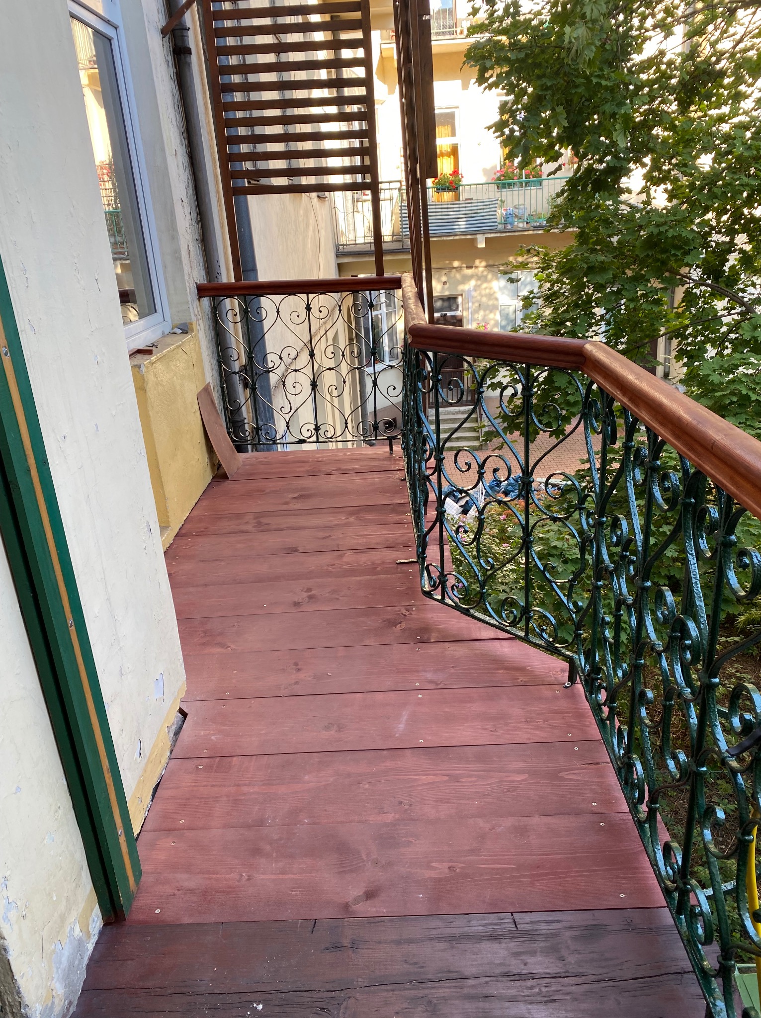 Czerwono-brązowy drewniany taras na balkonie z kutą, zieloną balustradą i drewnianą poręczą, widok z góry na zieleń ogrodu i budynki w tle.