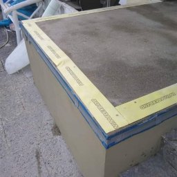 Best Tilers Group Kamil Pieczajka - Przygotowanie podłoża pod płytki: izolacja przeciwwodna narożnika z użyciem taśmy WIM FLEXBAND, widoczne łączenie ścian i podłogi, surowy beton w tle.