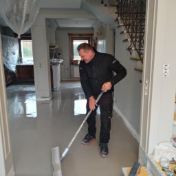 Best Tilers Group Kamil Pieczajka - Pracownik w czarnym ubraniu rozprowadza szarą wylewkę samopoziomującą na podłodze w domu, widoczne schody z zabezpieczonymi stopniami i narzędzia na schodach, pomieszczenie w trakcie remontu.