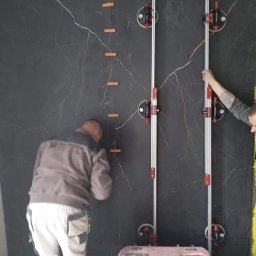 Best Tilers Group Kamil Pieczajka - Montaż wielkoformatowych płyt imitujących czarny marmur z jasnymi żyłami na ścianie, przy użyciu przyssawek i listew poziomujących, widoczne pomarańczowe kliny dystansowe.