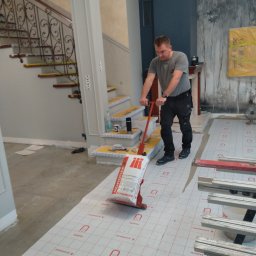 Best Tilers Group Kamil Pieczajka - Pracownik w trakcie układania podkładu podłogowego marki Schomburg w eleganckim wnętrzu z dekoracyjną ścianą i schodami z metalową balustradą, używając specjalistycznego wałka dociskowego.