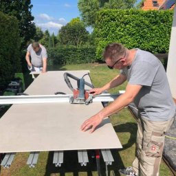 Best Tilers Group Kamil Pieczajka - Mężczyzna w okularach tnie płytę ceramiczną na zewnątrz za pomocą elektrycznej przecinarki, drugi mężczyzna przygotowuje kolejną płytę w tle, widoczne narzędzia i zielone krzewy.