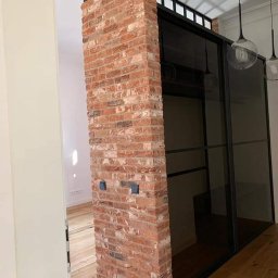 Best Tilers Group Kamil Pieczajka - Ceglana kolumna z dwoma gniazdkami elektrycznymi, oddzielająca przestrzeń z przesuwnymi, czarnymi drzwiami, widok na drewnianą podłogę.