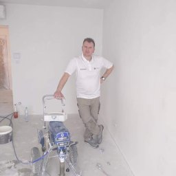 Best Tilers Group Kamil Pieczajka - Mężczyzna w białej koszulce polo i beżowych spodniach opiera się o maszynę do natryskowego malowania ścian w nowo pomalowanym, jasnym pomieszczeniu z widocznymi śladami farby na podłodze.