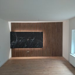 Best Tilers Group Kamil Pieczajka - Salon z drewnianą okładziną ścienną z podświetleniem LED i panelem z czarnego marmuru, jasna drewniana podłoga, grzejnik pod oknem.
