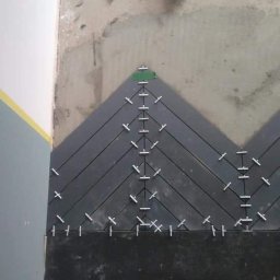 Best Tilers Group Kamil Pieczajka - Układanie ciemnych płytek podłogowych w jodełkę z użyciem krzyżyków dystansowych, widok z góry na fragment podłogi i buty osoby wykonującej prace.