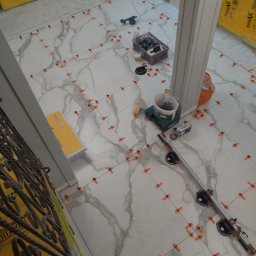 Best Tilers Group Kamil Pieczajka - Podczas układania płytek podłogowych z użyciem systemu poziomowania płytek, widoczne narzędzia glazurnicze, w tym przyssawki, poziomica i wiadro z klejem.