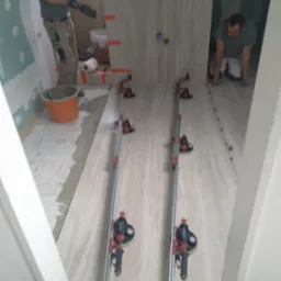 Montaż beżowych płytek imitujących marmur na podłodze i ścianie łazienki w trakcie remontu, widoczne narzędzia do cięcia płytek i poziomowania, pracownicy w trakcie układania płytek.