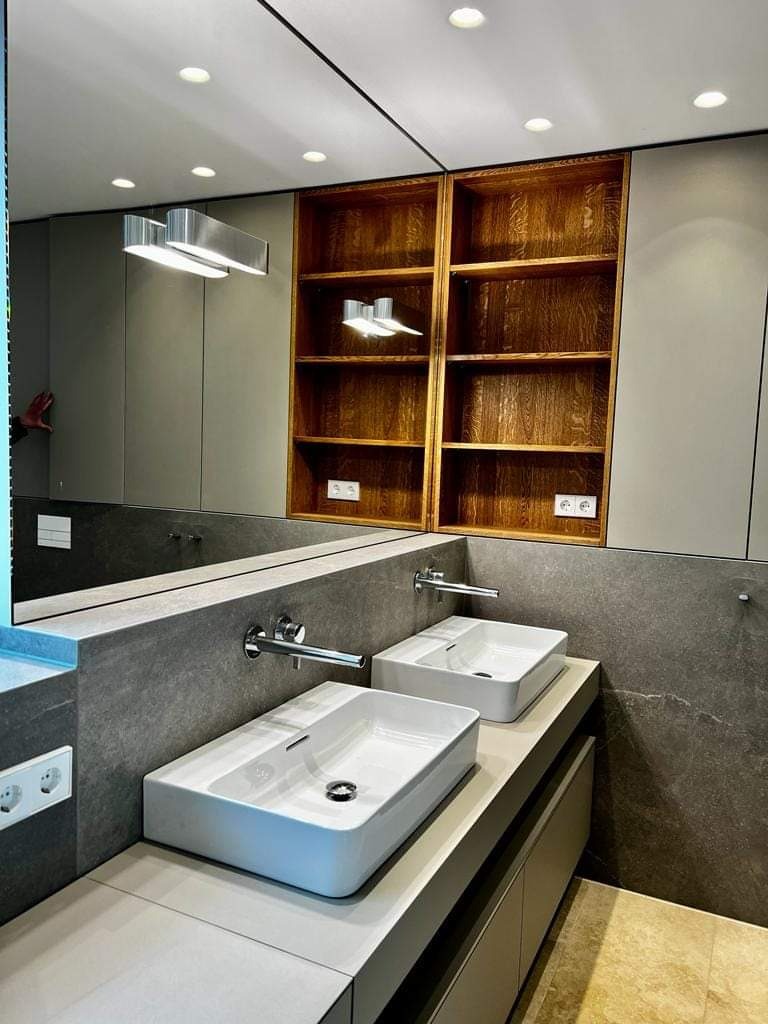 Nowoczesna łazienka z dwiema prostokątnymi umywalkami nablatowymi, chromowanymi bateriami ściennymi, szarą kamienną ścianą i drewnianymi półkami wnękowymi, oświetlona punktowymi lampami sufitowymi.