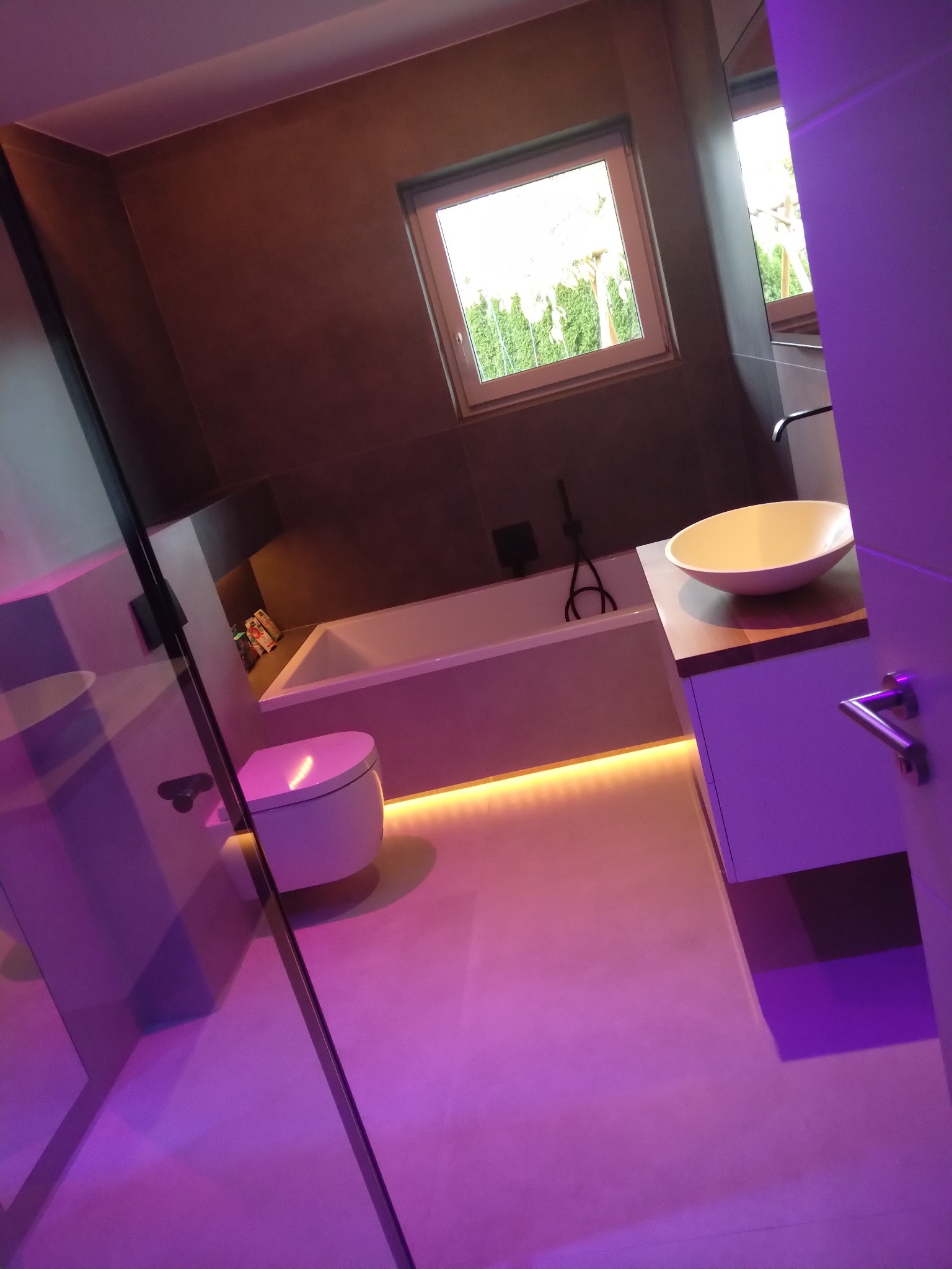 Nowoczesna łazienka z podświetleniem LED pod wanną, toaletą i szafką umywalkową, widok przez szklaną kabinę prysznicową, ciemne płytki na ścianach.