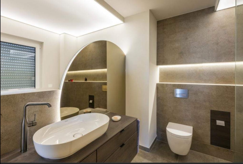 Nowoczesna łazienka z owalną umywalką nablatową, podświetlanym lustrem w kształcie łuku i ukrytą spłuczką WC w szarej, kamiennej okładzinie.