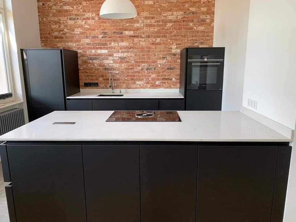 Nowoczesna kuchnia z wyspą: czarne szafki, biały blat z wbudowaną płytą grzewczą, ściana z czerwonej cegły i minimalistyczna lampa wisząca.