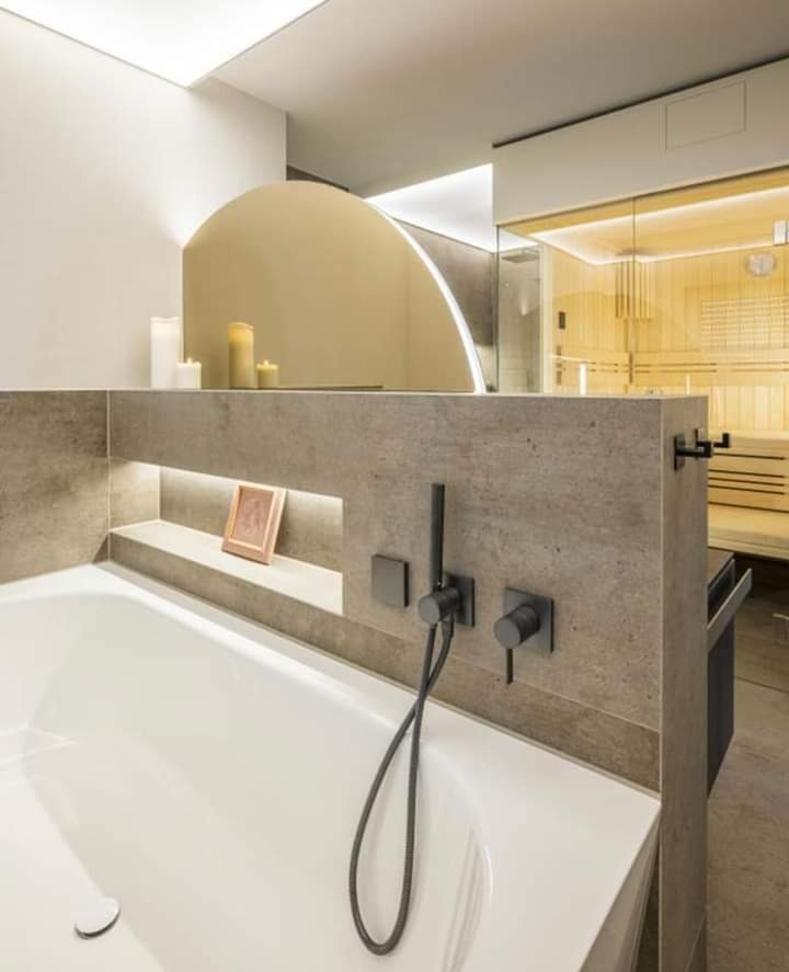 Nowoczesna łazienka z wanną, czarną armaturą i sauną w tle, oświetlona subtelnym światłem świec i LED, z dekoracyjną półką z ramką na zdjęcia.