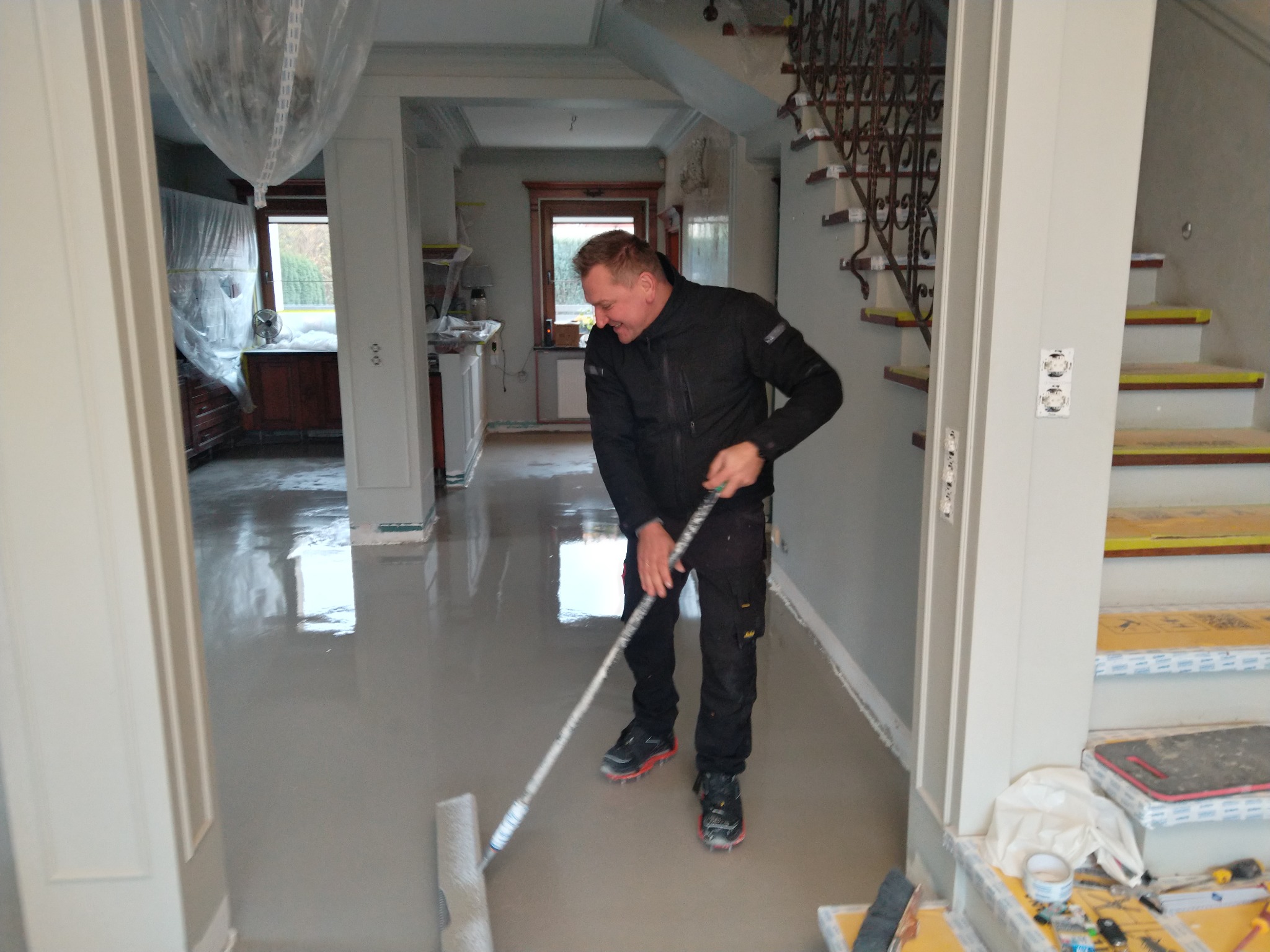 Pracownik w czarnym ubraniu rozprowadza szarą wylewkę samopoziomującą na podłodze w domu, widoczne schody z zabezpieczonymi stopniami i narzędzia na schodach, pomieszczenie w trakcie remontu.