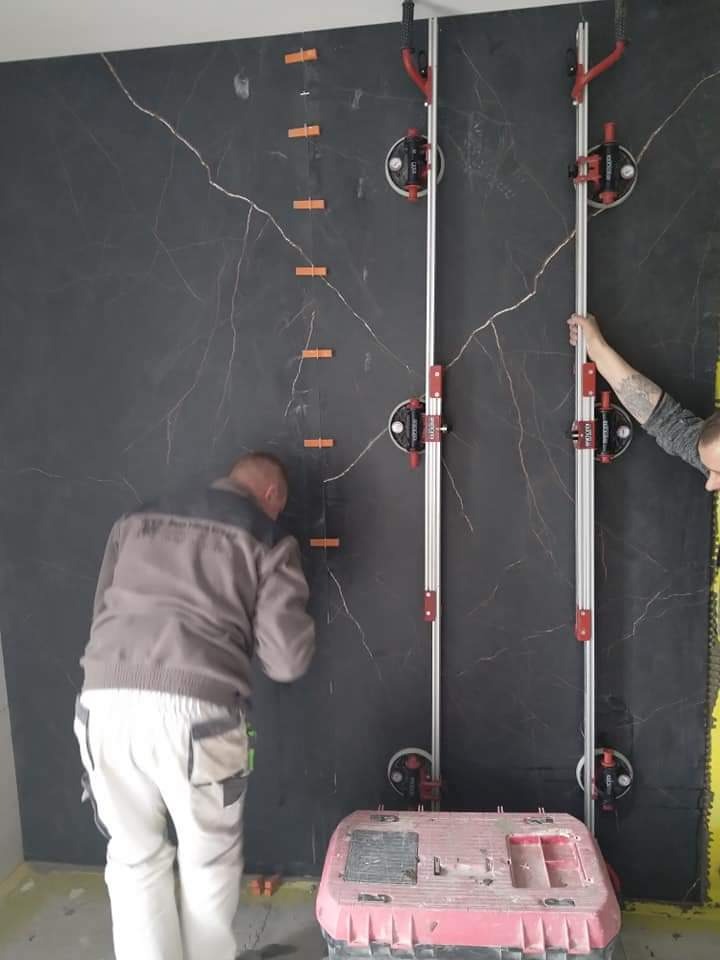 Montaż wielkoformatowych płyt imitujących czarny marmur z jasnymi żyłami na ścianie, przy użyciu przyssawek i listew poziomujących, widoczne pomarańczowe kliny dystansowe.