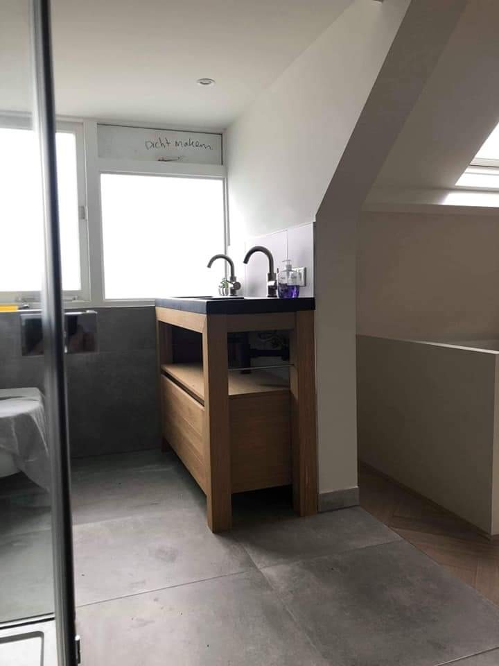 Nowoczesna łazienka na poddaszu z drewnianą szafką umywalkową z czarnym blatem i dwoma czarnymi kranami, szara podłoga i częściowo widoczna kabina prysznicowa, na oknie napis 'dicht maken'.