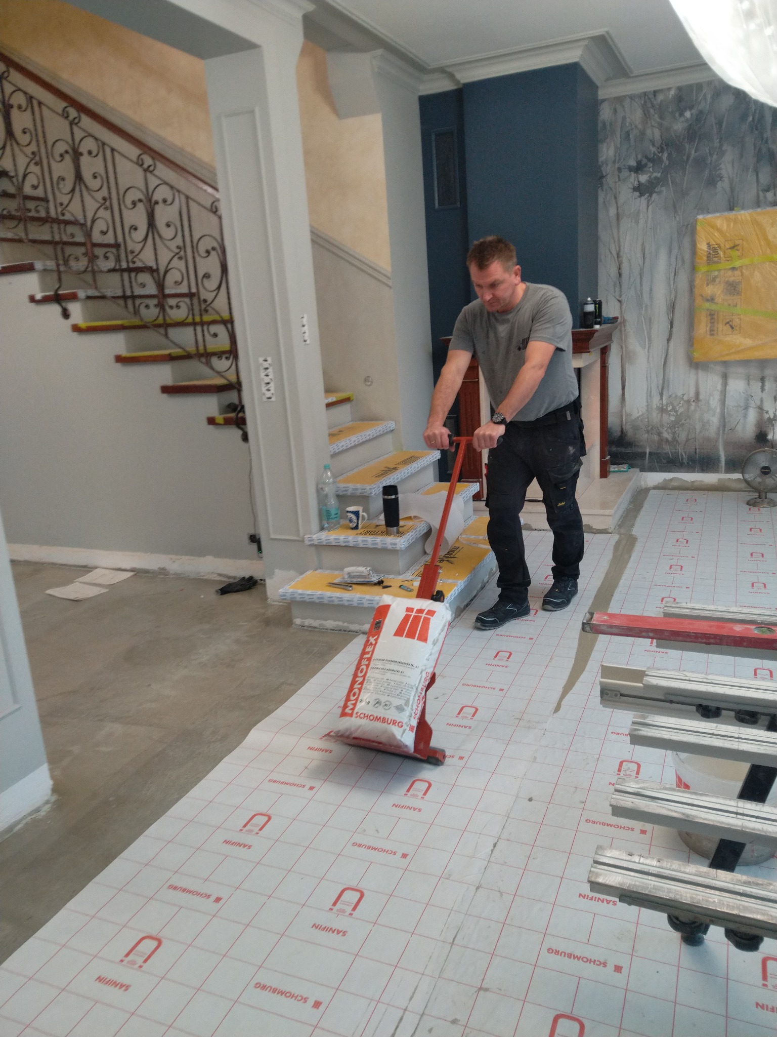 Pracownik w trakcie układania podkładu podłogowego marki Schomburg w eleganckim wnętrzu z dekoracyjną ścianą i schodami z metalową balustradą, używając specjalistycznego wałka dociskowego.