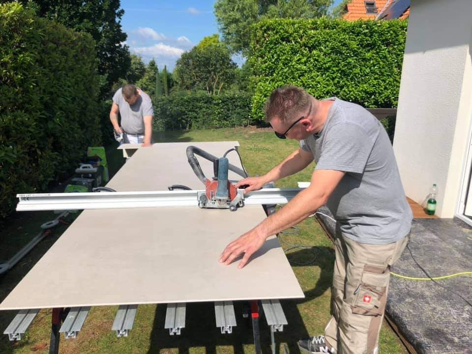 Mężczyzna w okularach tnie płytę ceramiczną na zewnątrz za pomocą elektrycznej przecinarki, drugi mężczyzna przygotowuje kolejną płytę w tle, widoczne narzędzia i zielone krzewy.