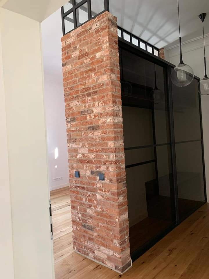 Ceglana kolumna z dwoma gniazdkami elektrycznymi, oddzielająca przestrzeń z przesuwnymi, czarnymi drzwiami, widok na drewnianą podłogę.