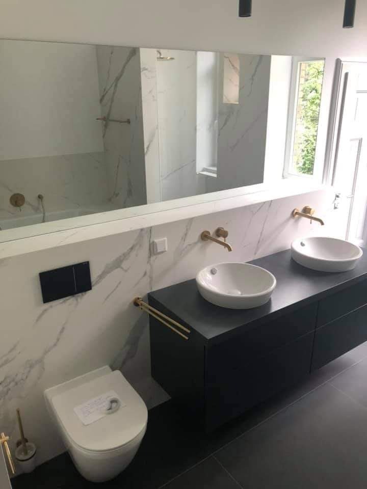 Nowoczesna łazienka z marmurowymi płytkami, podwójną umywalką nablatową, czarną szafką pod umywalką, złotymi bateriami i akcesoriami, oraz wiszącą toaletą z naklejką na desce.