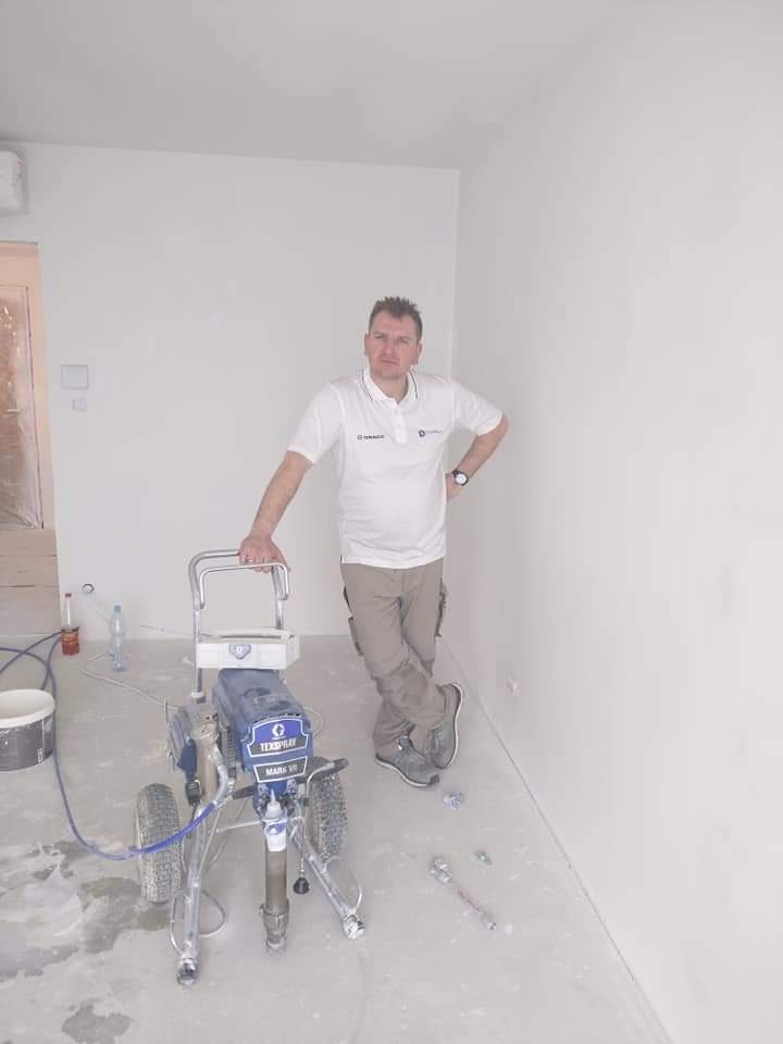 Mężczyzna w białej koszulce polo i beżowych spodniach opiera się o maszynę do natryskowego malowania ścian w nowo pomalowanym, jasnym pomieszczeniu z widocznymi śladami farby na podłodze.