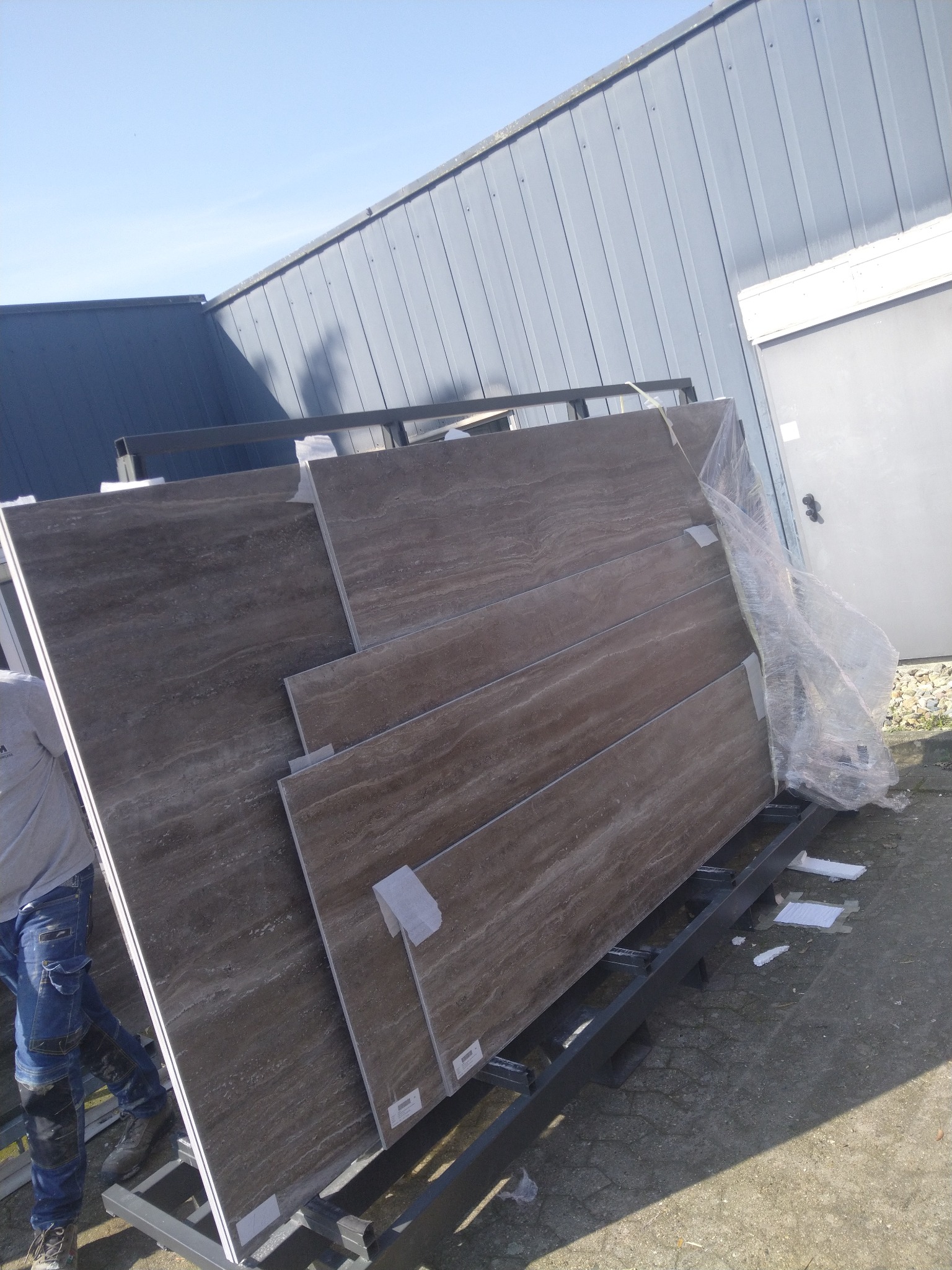 Duże, brązowe płytki imitujące trawertyn, zabezpieczone folią i umieszczone na metalowym stojaku, czekają na transport.