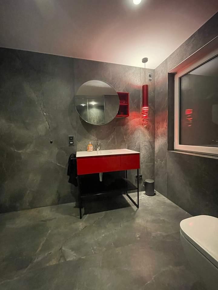 Nowoczesna łazienka z szarymi płytkami imitującymi marmur, czerwona szafka pod umywalką z okrągłym lustrem, wisząca czerwona lampa i biała toaleta.