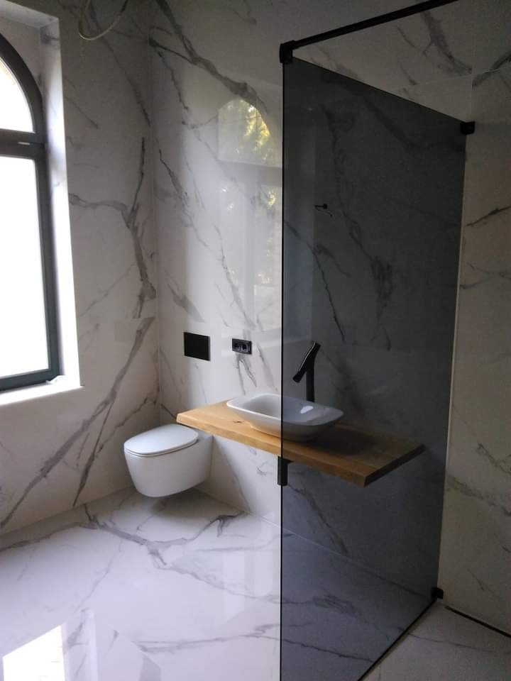 Nowoczesna łazienka z białymi marmurowymi płytkami, wiszącą toaletą, umywalką na drewnianym blacie i kabiną prysznicową z czarnymi profilami.