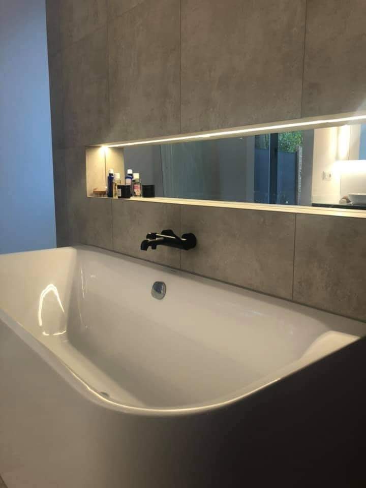 Nowoczesna łazienka z białą wanną, czarną armaturą i podświetlaną wnęką z kosmetykami, wykończona płytami w odcieniach szarości.