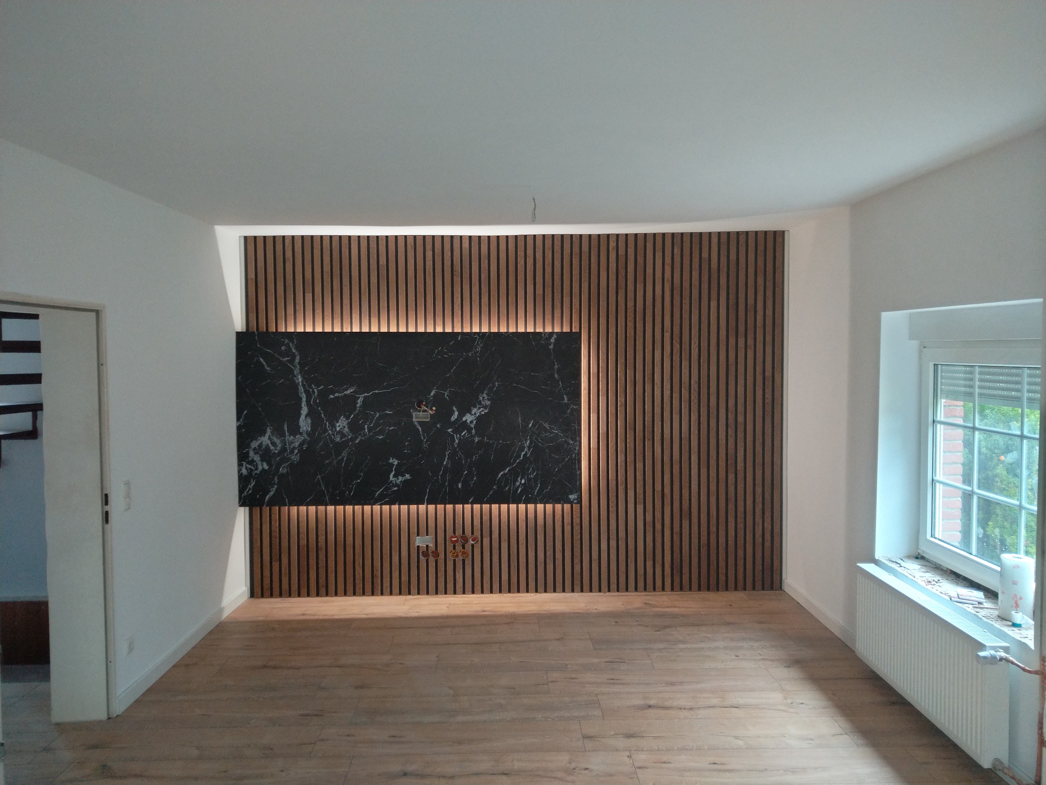 Salon z drewnianą okładziną ścienną z podświetleniem LED i panelem z czarnego marmuru, jasna drewniana podłoga, grzejnik pod oknem.