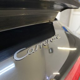 RB MOTORS RAFAŁ BOGACKI - Błyszczący, czarny Porsche Carrera 2 z chromowanym napisem, widoczne odbicia światła na lakierze, fragment tylnej lampy i aplikator do polerowania lakieru.