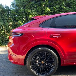 RB MOTORS RAFAŁ BOGACKI - Czerwony samochód typu SUV, prawdopodobnie Porsche Cayenne, z czarnymi felgami, zaparkowany na brukowanej powierzchni w Gdańsku, z zielonymi krzewami w tle. Widoczny fragment karoserii po detailing.