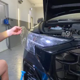 Nakładanie przezroczystej folii ochronnej na reflektor czarnego samochodu z otwartą maską, widoczne narzędzie do aplikacji i detale maskownicy
