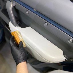 RB MOTORS RAFAŁ BOGACKI - Szczotkowanie jasnej tapicerki drzwi samochodowych, widoczna piana czyszcząca i ręka w czarnej rękawicy ochronnej.