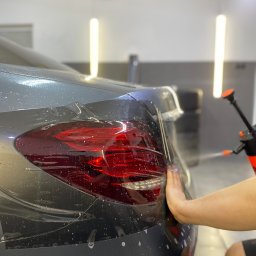 RB MOTORS RAFAŁ BOGACKI - Aplikacja folii ochronnej na tylną lampę samochodu w jasnym warsztacie, widoczne krople wody i dłoń pracownika wygładzająca folię. W tle butla ze spryskiwaczem.