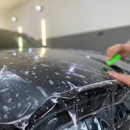 WrapConcept - Aplikacja folii ochronnej na czarny samochód w warsztacie, widoczne bąbelki wody i narzędzie aplikacyjne w dłoni.