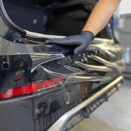 WrapConcept - Nakładanie folii ochronnej na tylny zderzak czarnego samochodu, widoczna ręka w czarnej rękawicy wygładzająca folię, otwarty bagażnik, krople wody na powierzchni.