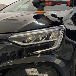 RB MOTORS RAFAŁ BOGACKI - Polerowanie lakieru czarnego samochodu za pomocą polerki Milwaukee Fuel, widoczny reflektor i fragment maski, odbicie osoby w lakierze.