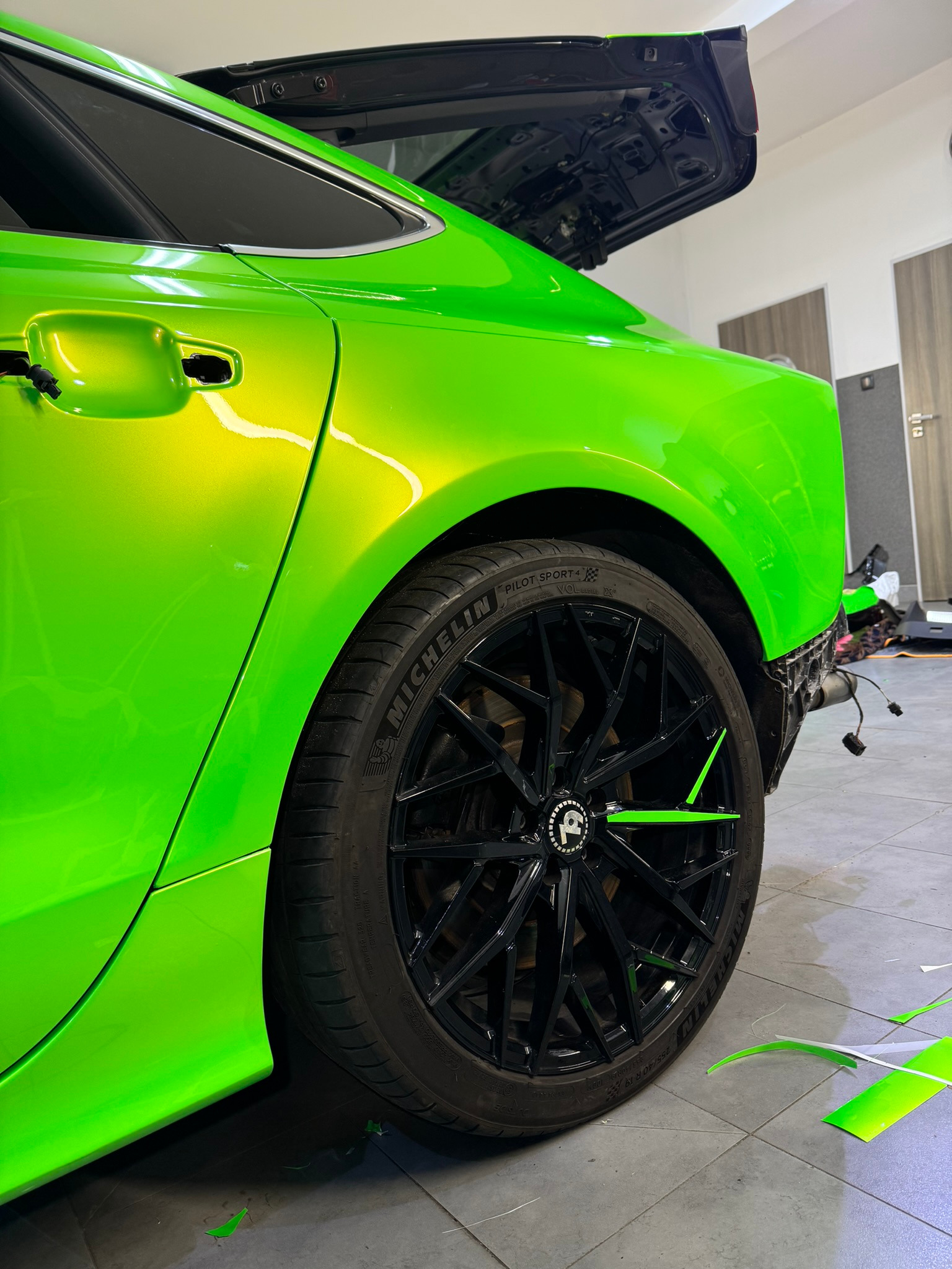 Jaskrawozielony samochód z otwartą klapą bagażnika, czarne felgi z zielonymi akcentami, widoczne ślady folii na podłodze, sugerujące proces oklejania.