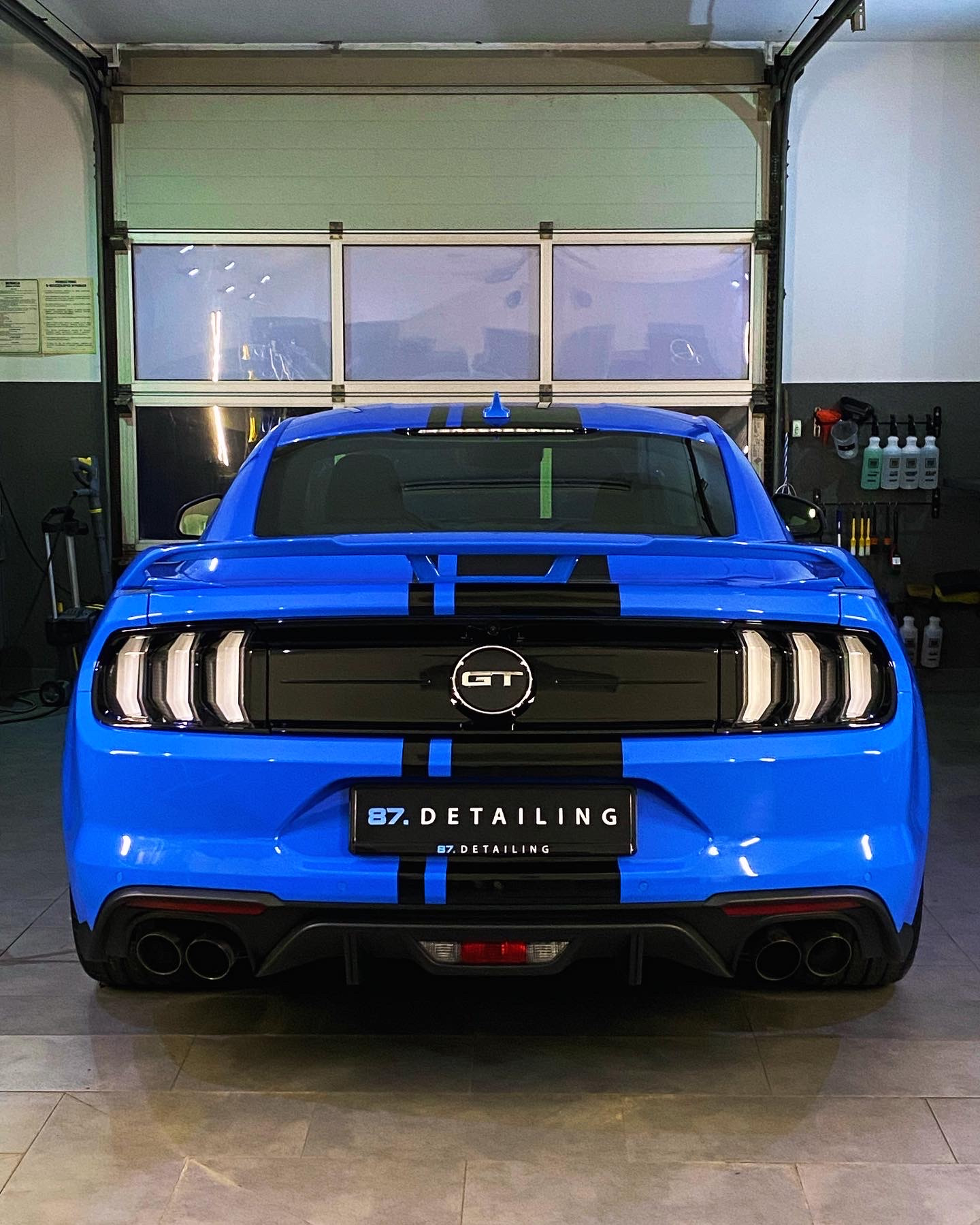 Błękitny Ford Mustang GT z czarnymi pasami, widok z tyłu, po detailingu w warsztacie, z widocznym logo firmy na tablicy rejestracyjnej.