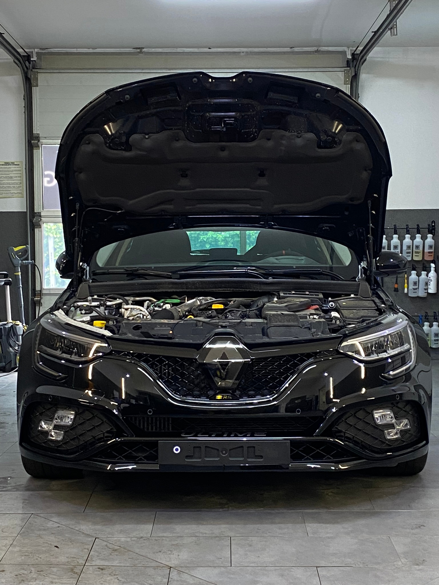 Czarny samochód z otwartą maską w garażu, widoczne elementy silnika, butelki z preparatami na ścianie w tle.