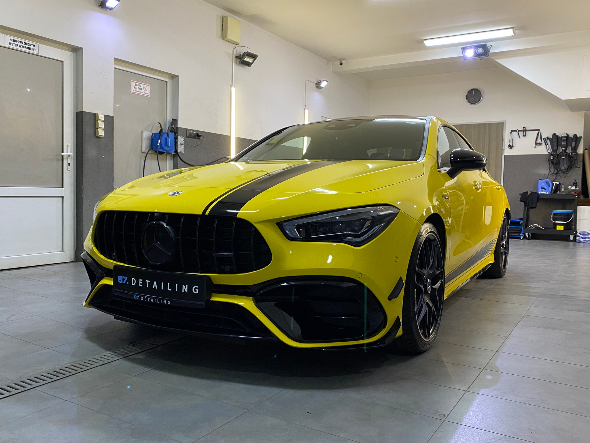 Żółty Mercedes CLA z czarnym pasem na masce, po detailingu, z widocznym logo firmy 87. Detailing na grillu, w jasnym garażu.